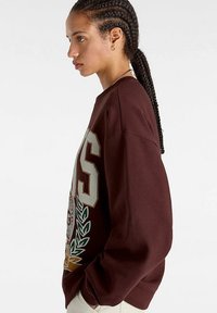 Bordeauxfärgad oversized sweatshirt med en grafisk design som har ett vapen och stora bokstäver. Tillverkad av ett mjukt, texturerat tyg.