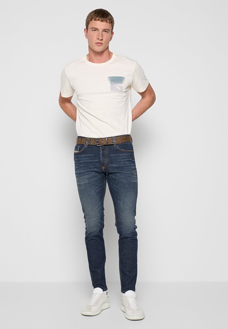 Junger Mann, der ein weißes T-Shirt mit einer Tasche, blaue Jeans, einen braunen Gürtel mit Löchern und weiße Sneakers trägt, steht mit den Händen hinter dem Rücken.
