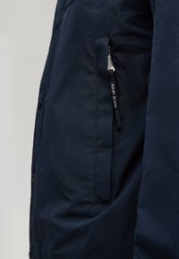 Granatowa kurtka z pionową kieszenią na zamek; suwak ozdobiony etykietą "JOOP! JEANS" w białym napisie na czarnym materiale.