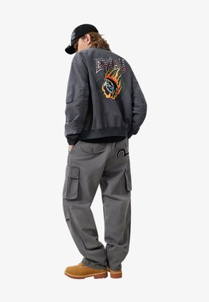 Persona con giacca bomber grigio scuro con design colorato di fiamme e testo, pantaloni cargo grigi, stivali color sabbia e berretto nero, in piedi con le mani in tasca.