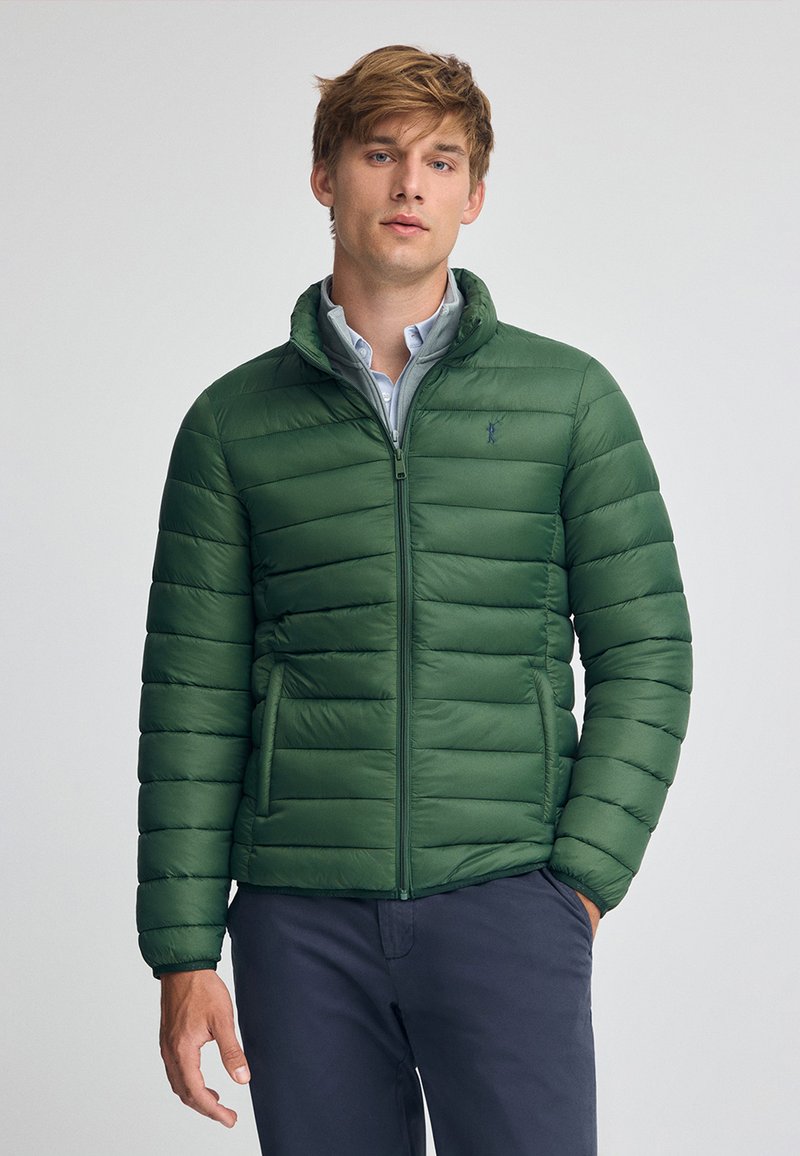 Grön vadderad jacka med horisontellt quiltat mönster, framdragkedja och två sidofickor. Bärs över en ljusblå skjorta med ståkrage.