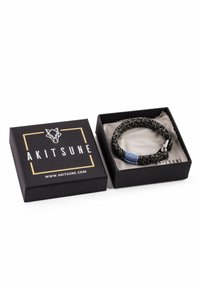 Bracelet tressé noir avec une bande bleue et un fermoir argenté dans une boîte noire à côté d'une boîte fermée étiquetée Akitsune.