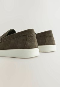 Chaussures marron texturées à enfiler avec semelles en caoutchouc blanches vues de dos sur un fond clair uni.