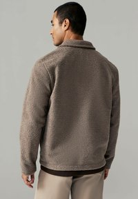 Geacă din fleece de culoare marron, cu o suprafață texturată, guler și mâneci lungi. Bază securizată și stratificată subtil cu un top marron dedesubt.