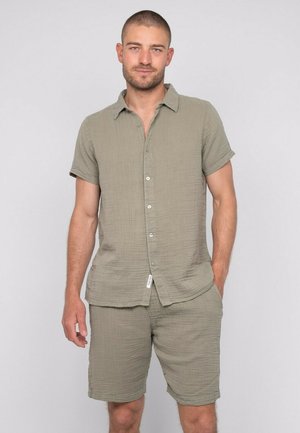 Homme aux cheveux courts portant une chemise boutonnée à manches courtes assortie couleur olive clair et un short, debout avec une main dans la poche.