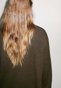 Brun, texturerad tröja med en lös passform, bärs av en person med långt, vågigt hår, sett bakifrån mot en ljus bakgrund.