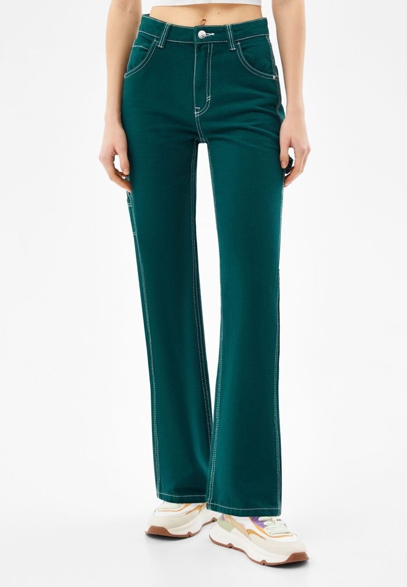 Nainen, joka on pukeutunut korkeavyötäröisiin smaragdinvihreisiin levenevästi leikatuihin housuihin, joissa on valkoinen kontrastivärinen tikkaus, sekä monivärisiin platform-kenkiin, seisoo valkoista taustaa vasten.