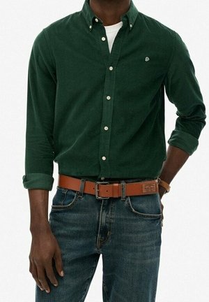 Homme portant une chemise à manches longues vert foncé avec les poignets retroussés, un jean bleu, une ceinture en cuir marron et une montre.