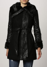 Cappotto nero con maniche in pelle trapuntata, colletto e polsini in pelliccia, vita con cintura, indossato con jeans blu strappati.