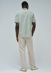 Camisa de manga corta de color verde claro con una textura sutil, combinada con pantalones beige holgados y espadrilles de color claro, vista desde atrás.