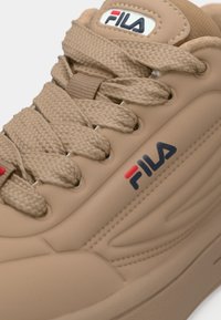 Beige sportsko med teksturert syntetisk materiale, med vevde snørebånd, en fremtredende Fila-logo i blått og rødt, og glatte detaljer.