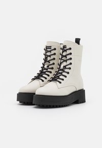 Botines de cuero blanco con cordones negros, suelas negras gruesas y una lengüeta en la parte trasera. Presentan una textura suave y una marca mínima.