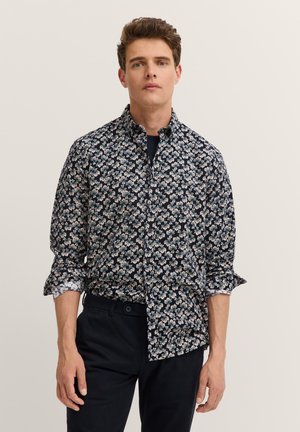 Junger Mann trägt ein dunkles, florales Hemd mit hochgekrempelten Ärmeln und schwarze Hose, steht vor einem schlichten hellen Hintergrund.