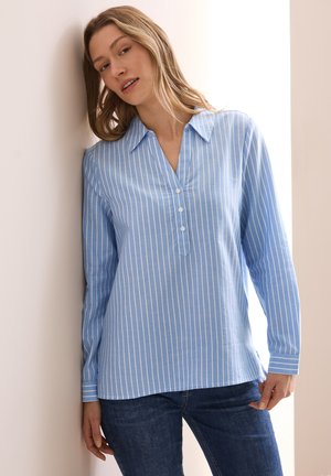 Camisa de manga larga azul claro con rayas blancas verticales, una tapeta de botones y un cuello. Combinada con jeans azul oscuro.