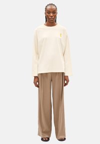 Langärmliges Baumwollhemd in Beige mit einem gelben Schleifenlogo, kombiniert mit weit geschnittenen, hellbraunen Hosen und schwarzen Slides.