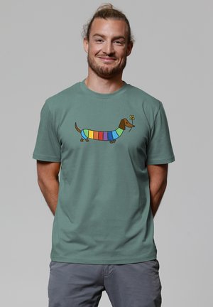 Homme barbu aux cheveux attachés portant un t-shirt vert avec un teckel rayé coloré tenant une fleur jaune dans sa bouche.