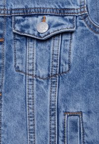 Helles blaues Denim-Gewebe mit sichtbaren Nähten; ausgestattet mit einem silbernen Druckknopf und einer Brusttasche. Die Textur wirkt weich und leicht ausgebleicht.