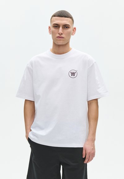 Jeune homme aux cheveux courts portant un T-shirt blanc avec un logo circulaire "W" sur la poitrine et un short noir, sur un fond uni.
