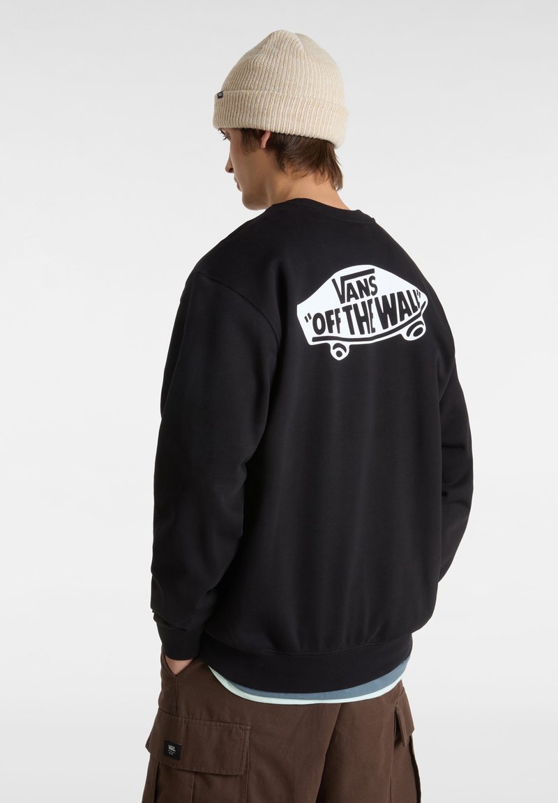 Sort sweatshirt med hvid "VANS OFF THE WALL" logo på ryggen, manchetterne og kanten er ribbede, båret over en lys blå skjorte og brune bukser.