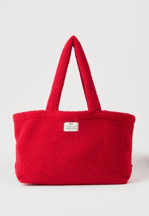 Sac fourre-tout rouge vif texturé avec deux poignées et une petite étiquette blanche rectangulaire à l’avant sur un fond uni.