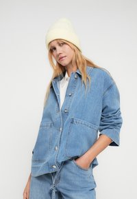 Ung kvinne iført gul strikket lue, lyseblå denimjakke med store lommer og matchende denim bukser, poserer med én hånd i lommen.