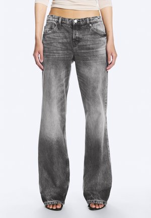 Personne portant un jean noir délavé taille haute à jambes larges avec cinq poches et des sandales noires ouvertes à l'avant, debout devant un fond uni.