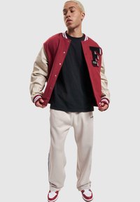 Veste varsity rouge et beige avec des lettres noires, associée à un t-shirt noir et un pantalon de survêtement beige avec des rayures sur les côtés. Baskets rouges.