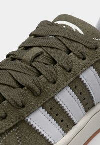 Zielone zamszowe sneakersy posiadają płaskie sznurówki, białe skórzane paski, perforacje na palcach i gumową podeszwę z niewielkim podniesieniem.