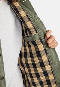 Chaqueta de tela verde con un forro interior de cuadros que presenta patrones amarillos y marinos. La chaqueta está parcialmente desabrochada, mostrando el diseño interior.