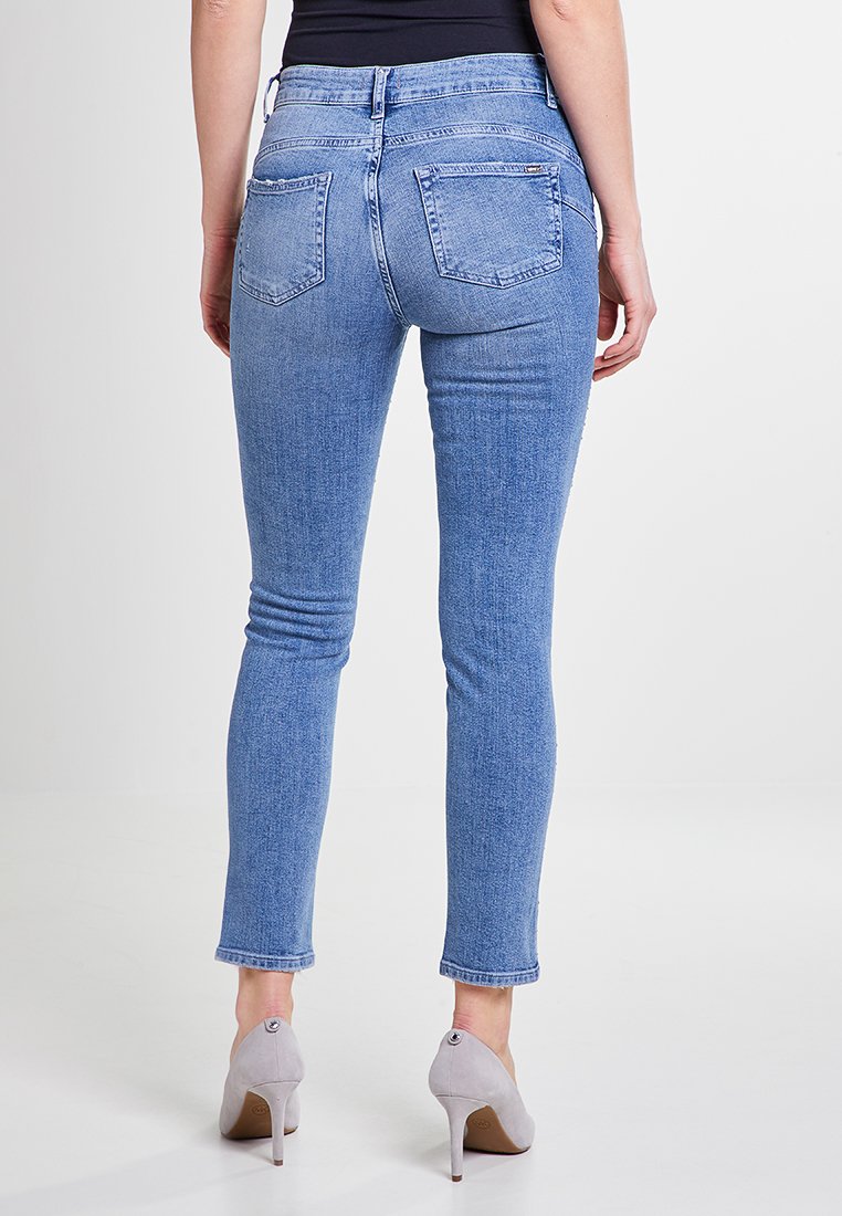 Donna che indossa jeans skinny azzurri chiari e tacchi alti grigi, in piedi di spalle contro uno sfondo bianco semplice.