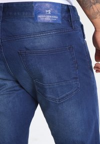 Jeans de mezclilla azul oscuro con una etiqueta de cuero azul que lleva la marca "Scotch & Soda", y un bolsillo trasero con detalles de costura.