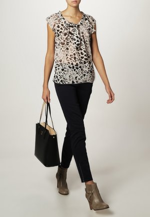 Vrouw loopt, draagt een blouse met luipaardprint, zwarte smalle broek, taupekleurige enkellaarzen en draagt een zwarte tote tas.