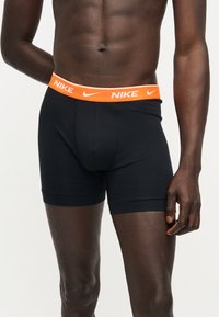 Αρσενικός κορμός που φοράει μαύρα boxer briefs Nike με πορτοκαλί ζώνη μέσης με λευκά λογότυπα Nike, στέκεται σε απλό φωτεινό φόντο.