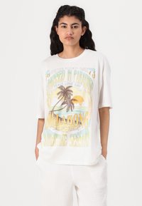 T-shirt bianco oversize con stampa grafica colorata di un tramonto, palme e testo. Vestibilità rilassata con maniche corte e scollatura rotonda.