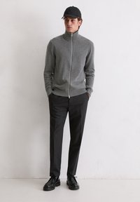 Grauer Reißverschluss-Pullover aus weichem Stoff, kombiniert mit dunklen, maßgeschneiderten Hosen und schwarzen Schnürschuhen. Eine schwarze Mütze rundet das Outfit ab.