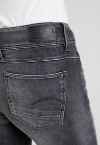 Szare dżinsy denimowe mają skórzaną łatę z logo, widoczne szwy oraz tylnią kieszeń o zaokrąglonym kształcie. Klasyczny styl pięciu kieszeni.