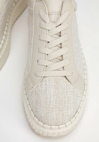 Beige teniska s texturovaným svrškem, odpovídajícími tkaničkami, hladkými koženými akcenty a silnou gumovou podrážkou na bílém pozadí.