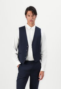 Marineblaue Weste mit einem taillierten Design, vorderen Knöpfen, zwei Taschen, getragen über einem weißen Hemd mit Knopfleiste, was einen formellen Eindruck vermittelt.