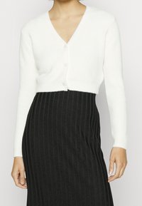 Weißer, verkürzter Cardigan aus strukturiertem Stoff mit V-Ausschnitt und drei Knöpfen, kombiniert mit einem schwarzen, gerippten Rock.