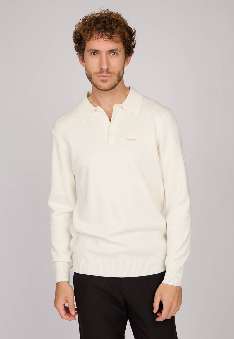 Polo shirt blanc à manches longues avec un col pointu, des poignets côtelés et une coupe slim. Présente un logo doré subtil sur la poitrine.