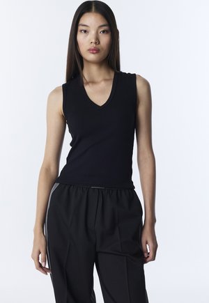 Jonge vrouw met lang, recht haar, gekleed in een zwarte mouwloze top met V-hals en loszittende zwarte broek, staand tegen een eenvoudige witte achtergrond.