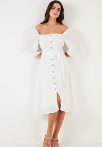 Bílá lněná šaty s off-shoulder nafouklými rukávy, předními knoflíky, vázacím páskem v pase a bočním rozparkem pro k dodatkový detail.