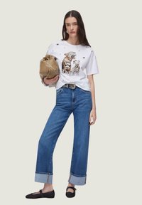 Bijela majica s grafikom jaguara, u kombinaciji sa širokim trapericama na preklop. Drži bež torbu, nosi crne ravne cipele.