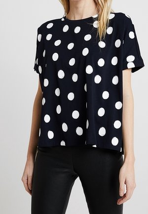 Kobieta w czarnej koszulce z krótkim rękawem w duże białe grochy i czarnych spodniach, stojąca na jednolitym jasnym tle.