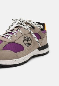 Baskets beige et violettes avec lacets à motif noir et blanc, semelle blanche tachetée, languette orange au talon et logo d'arbre noir sur le côté.