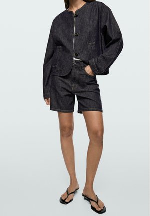 Donna con giacca di jeans scuro abbottonata e pantaloncini abbinati, indossa infradito nere e sta in piedi con una mano in tasca.