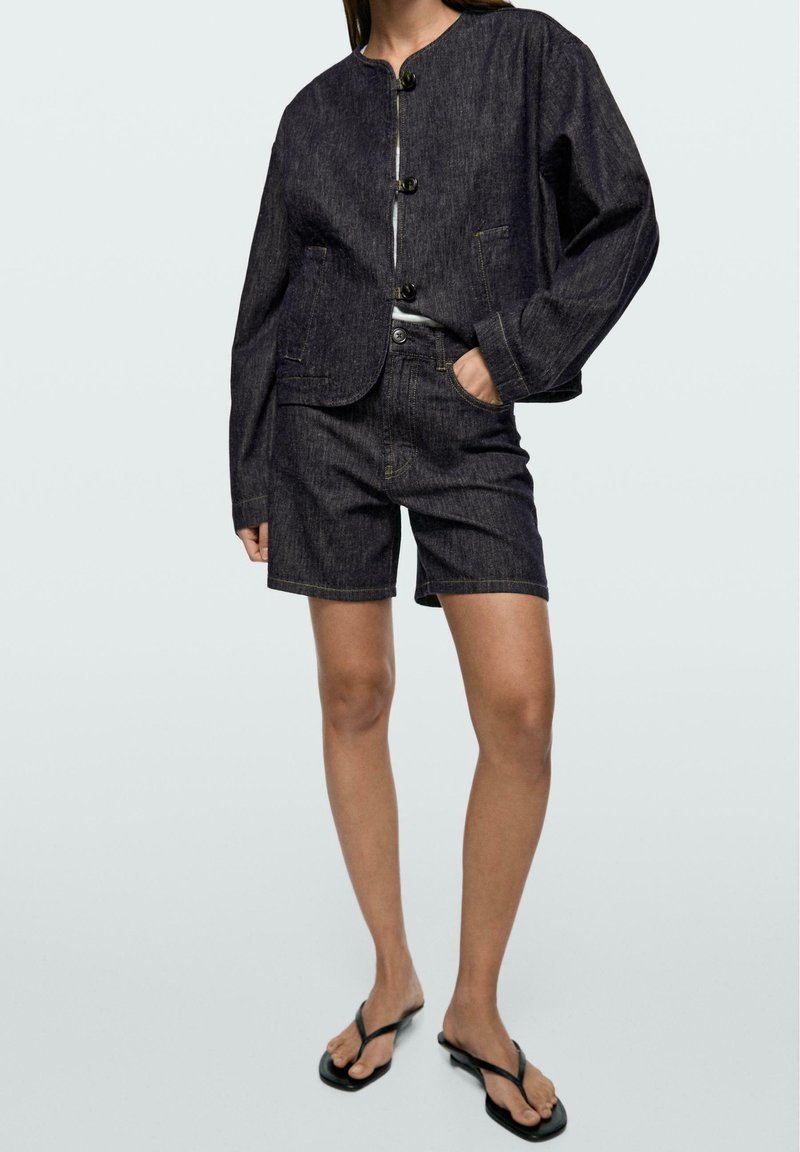 Kobieta w ciemnej, dżinsowej, zapinanej na guziki kurtce i dopasowanych szortach, w czarnych klapkach, stojąca z jedną ręką w kieszeni.