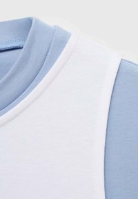 Close-up van gelaagde kleding met een witte mouwloze top over een lichtblauw shirt met lange mouwen en een geribbelde kraagdetail.