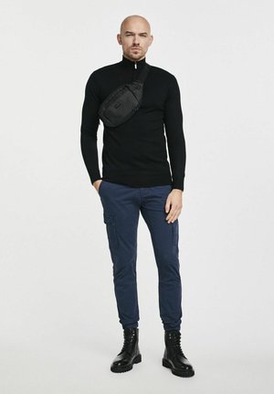 Czarny sweter z dzianiny, granatowe spodnie cargo, czarne buty bojowe i czarna torba na przekątnej. Czyste linie, dopasowana sylwetka.