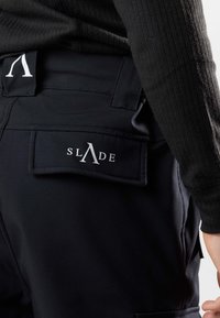 Czarne spodnie o teksturowanym wykończeniu, z tylną kieszenią z białym logo "SLADE" i gładkim paskiem w talii.